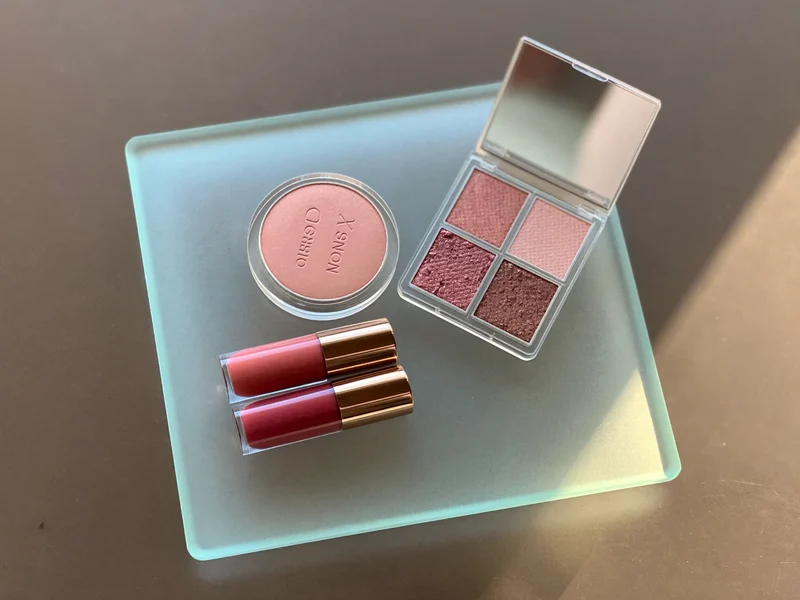 Rouge, zwei Lippenstifte und eine Lidschattenpalette in gedämpften Tönen auf einem Glastablett