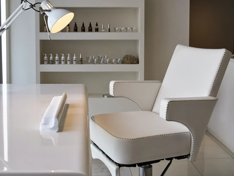 Aufgeräumter Behandlungsraum mit Maniküretisch, kleiner Lampe, leerem Stuhl und Regal voller Flaschen